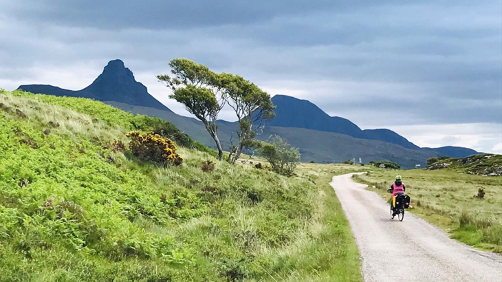 A winding path with green grass either side, dark mountains in the distance and a woman cycling away from the camera wearing high-vis and a helmet on an overcast day in Scotland's Highlands
