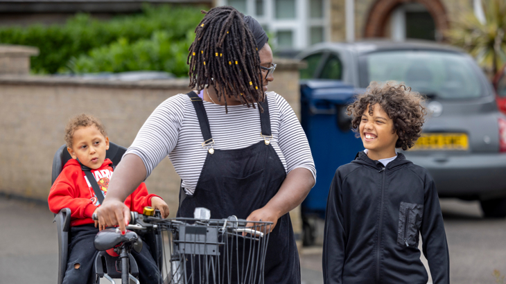 A woman with dreadlocks and black dungarees pushing a bike with a child sat in a child's seat on the bike while smiling at a young boy walking alongside her