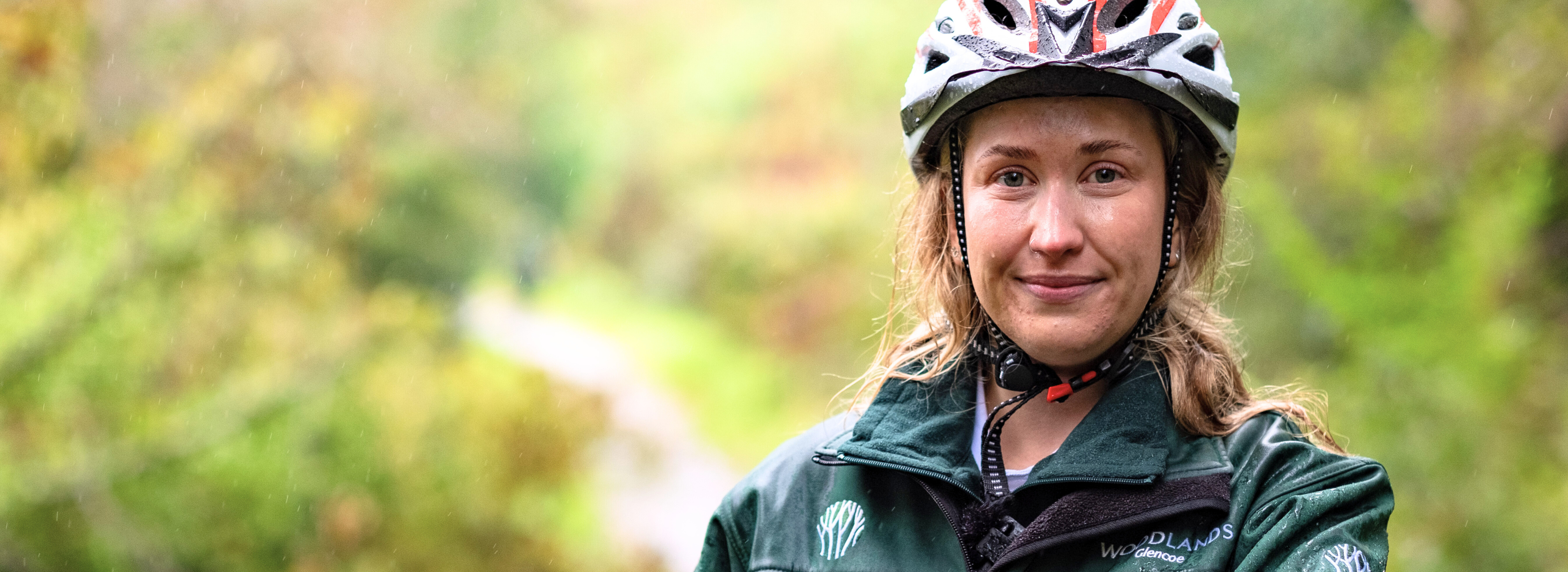A woman wearing a cycle helmet is out on the Network, in the rain. She has stopped and is looking directly at the camera, smiling slightly.