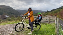 A mother stands astride her e-bike with her young son sat on the back, with a view of a town in a valley in the background.