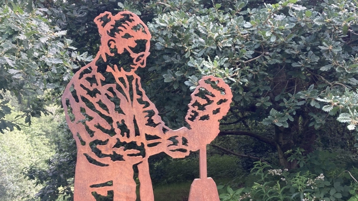 A steel sculpture of a woman with glasses and wearing her hair in a bun works on a sculpture surrounded by lush green trees and grass.