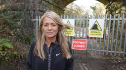 A person with long blonde hair smiling in front of a path that has been close off. Behind them is a sign that reads "footway closed".