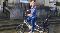 An adult woman stood astride a silver e-bike in front of a civic building in Swansea.