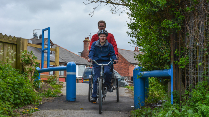 Two people riding a tandem trike through a wide barrier at the start of a Network path in a residential area